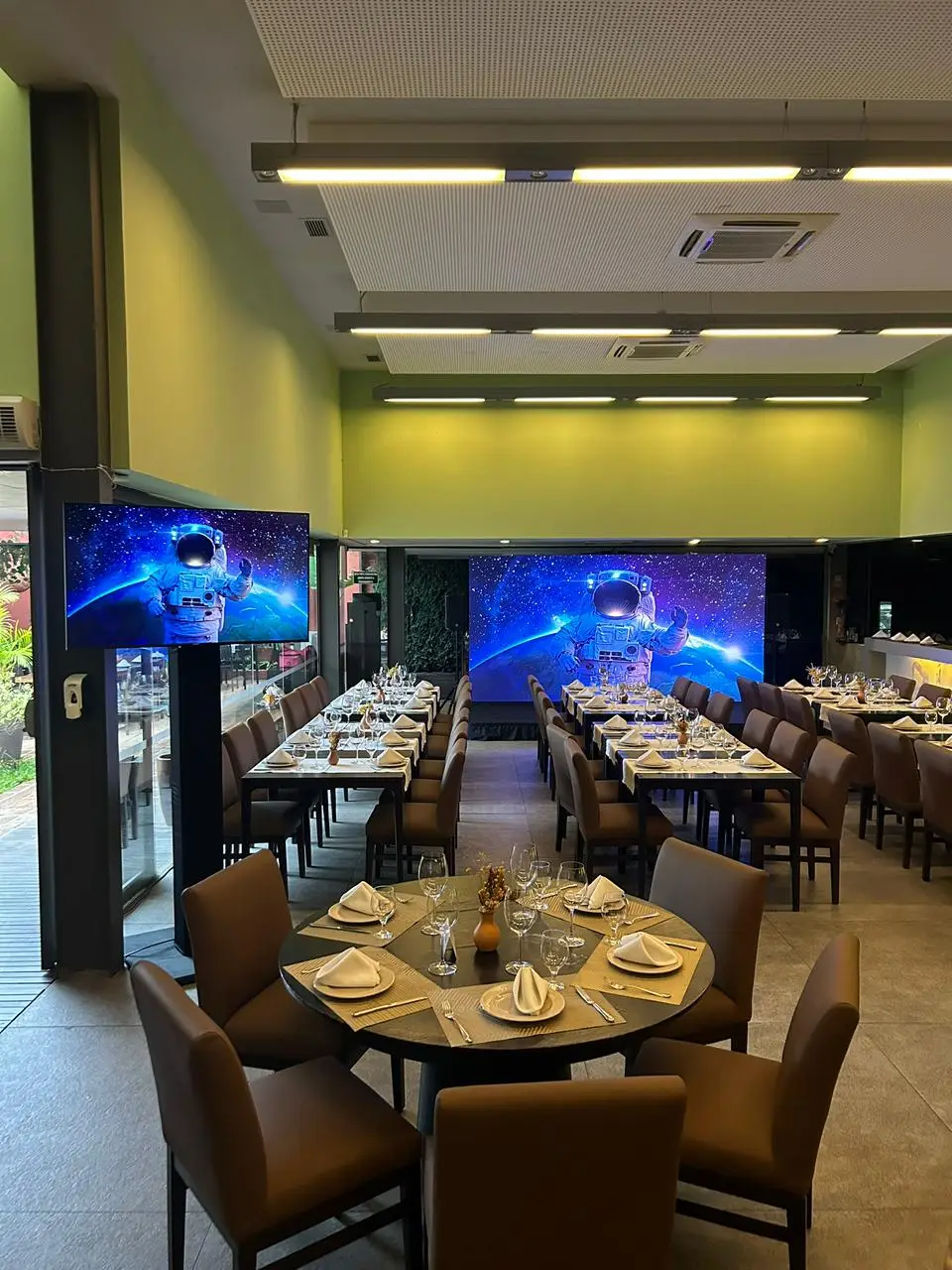 Salão preparado para jantar corporativo com mesas postas, painel de LED ao fundo e um monitor em pedestal, ambos exibindo a mesma arte de astronauta.