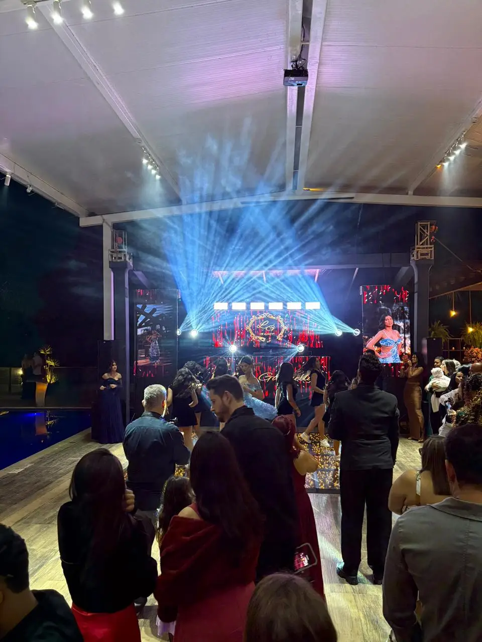 Pista de LED iluminada em piso elevado durante a entrada de uma festa de 15 anos, com convidados em volta e feixes de luz azul acima.