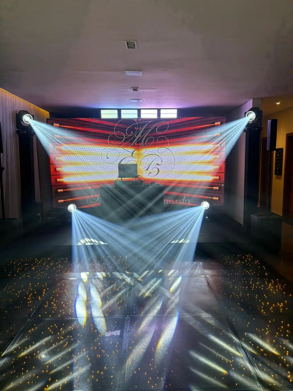 Pista de LED preta com pontos de luz dourados espalhados pelo piso, recebendo feixes brancos de quatro refletores móveis, com painel de LED ao fundo.