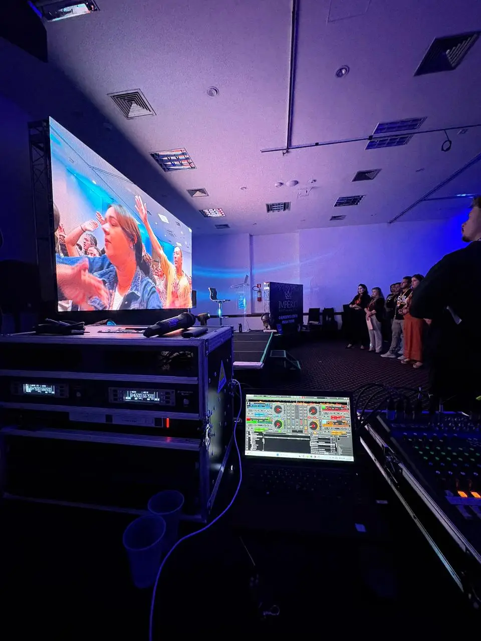 Mesa de operação durante um evento, com racks de receptores de microfone sem fio, microfones de mão, notebook e mesa de som, em frente a um painel de LED aceso.