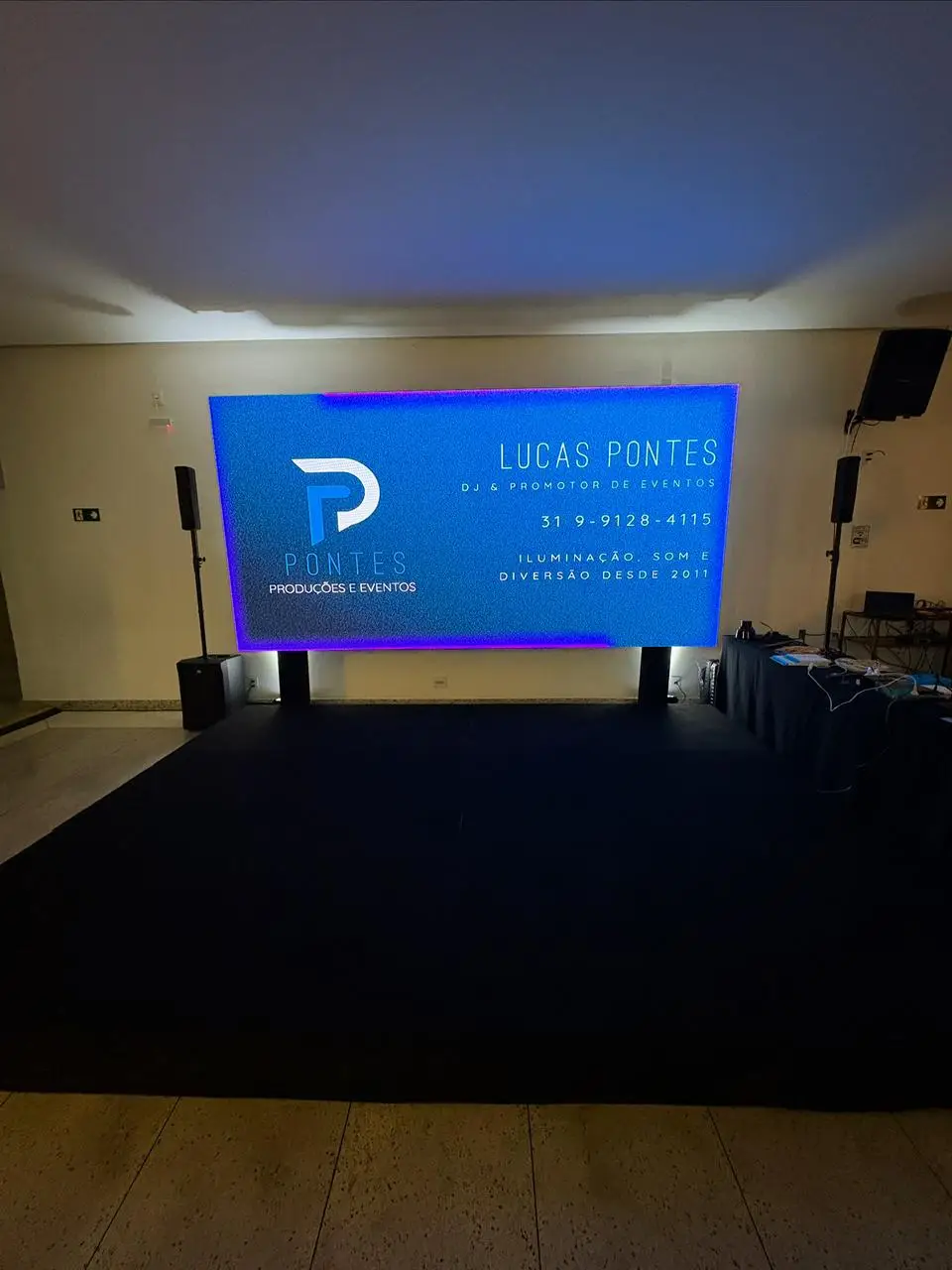Palco baixo forrado de preto em salão, sustentando um painel de LED sobre bases, com caixas de som em tripé nas laterais.