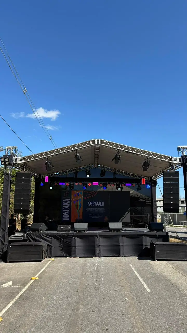 Palco ao ar livre com cobertura de treliça metálica, caixas de som line array penduradas nas torres laterais, refletores fixados na estrutura e painel de LED ao fundo do palco.