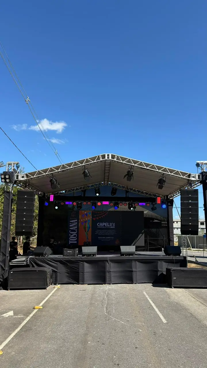 Palco coberto por estrutura de treliça em evento ao ar livre, com caixas de som line array penduradas nos dois lados, monitores de retorno na frente do palco e painel de LED ao fundo.