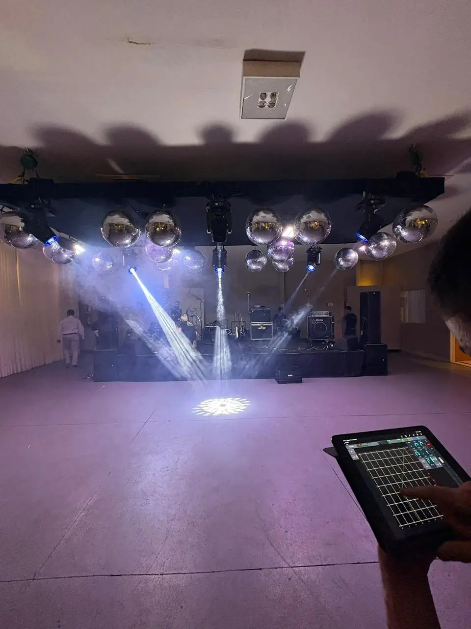 Técnico segura um tablet com o aplicativo de uma mesa de som digital aberto na tela de equalização, diante de um palco com bateria, amplificadores e refletores acesos.