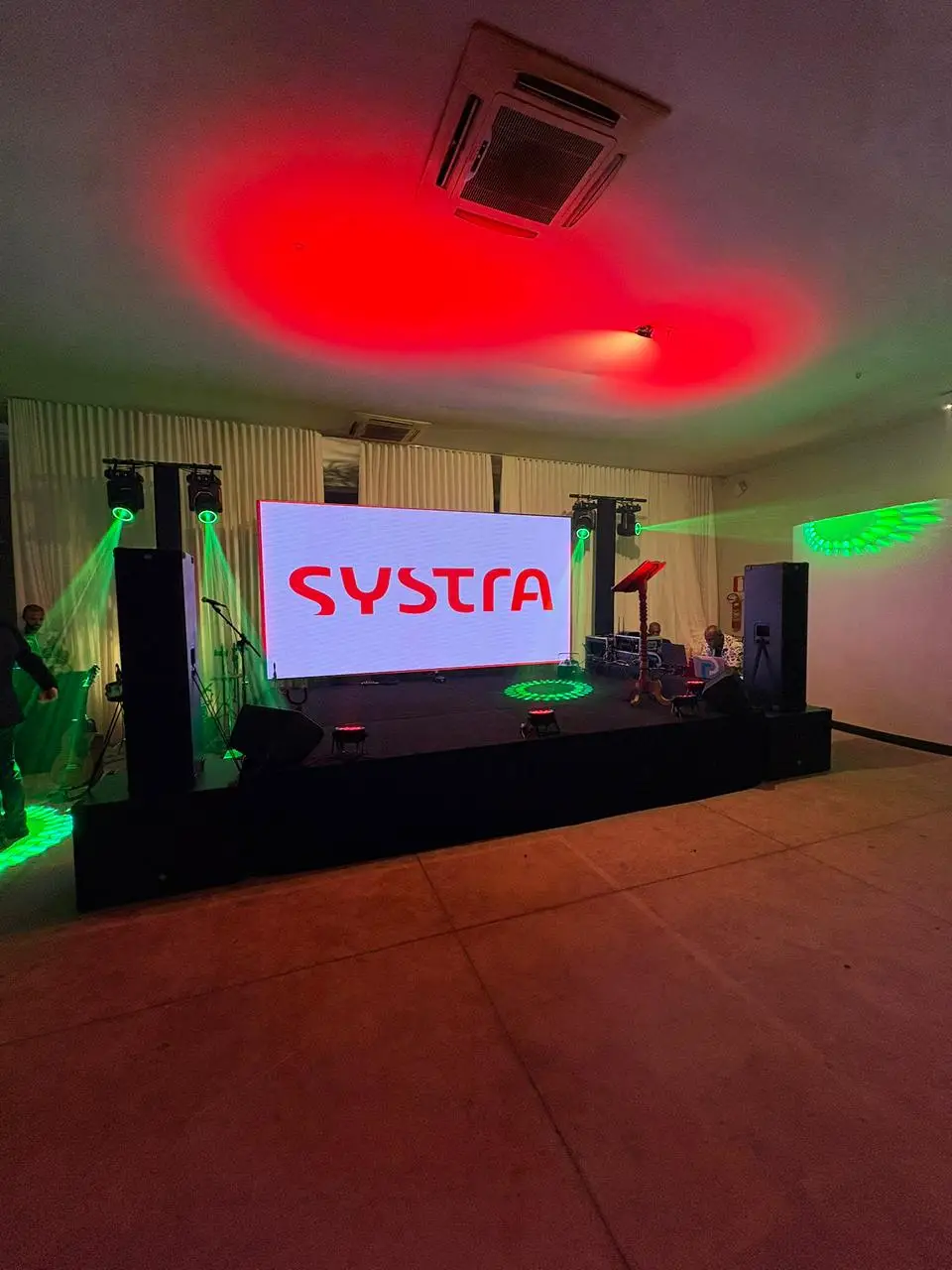 Painel de LED exibindo a marca Systra sobre um palco em salão fechado, com refletores móveis lançando feixes verdes e caixas de som nas laterais.